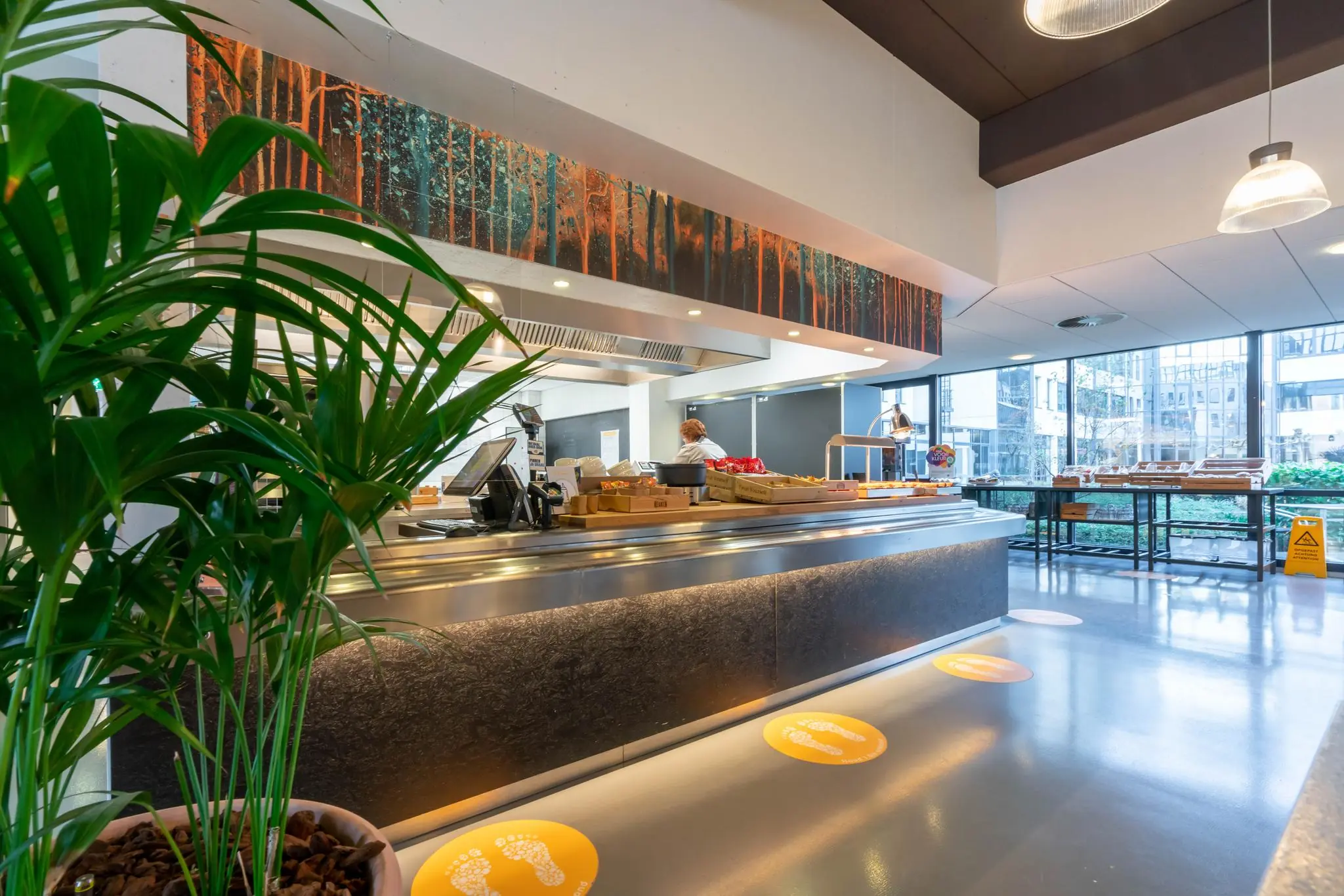 Moderne bedrijfskantine aan de De Bruyn Kopsstraat met een uitgiftebalie, lunchproducten en grote ramen met uitzicht op een binnentuin.